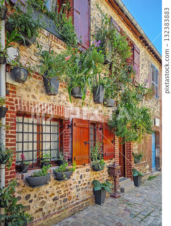 A quaint old masonry building with wooden shutters, adorned with numerous planters overflowing with greenery and flowers hanging on the facade. Rustic cobblestone path runs along the base of the house 132383883