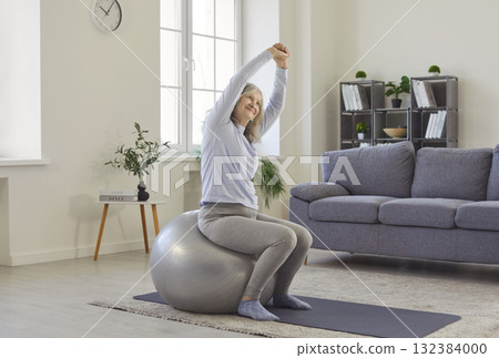 Old grey haired woman exercising with fitball, enjoying gymnastics at home, keeps arms above head 132384000
