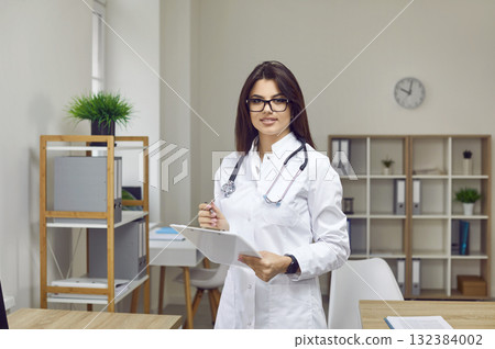Portrait of young woman doctor in white coat standing in staff room and holding clipboard 132384002