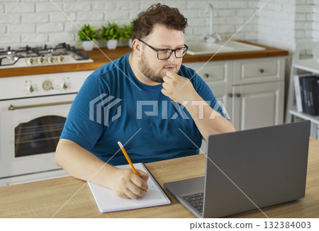 Plus size man studying online at home kitchen with laptop, notebook and pencil Plus size man studying online at home kitchen with laptop, notebook and pencil 132384003