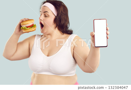 Overweight woman eats burger, uses calorie tracker and shows her mockup mobile phone 132384047