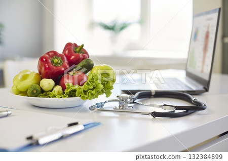 Plate of vegetables and stethoscope on clinic desk near laptop screen, daylight Plate of vegetables and stethoscope on clinic desk near laptop screen, daylight 132384899