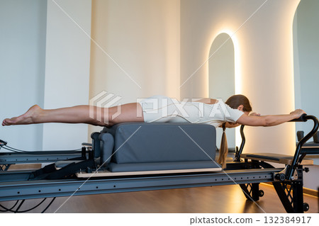 Woman doing Swan exercise on Pilates Reformer for the chest and back muscles, focusing on flexibility, strength, and control with determination Woman doing Swan exercise on Pilates Reformer for the chest and back muscles, focusing on flexibility, strength, and control with determination 132384917