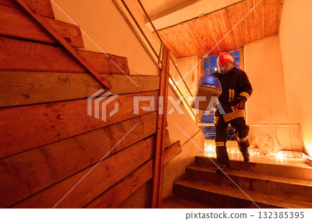 Brave firefighter carrying a rescued cat in a portable cage down smoky stairs during a building fire, symbolizing courage, compassion, and dedication to saving both human and animal lives in 132385395