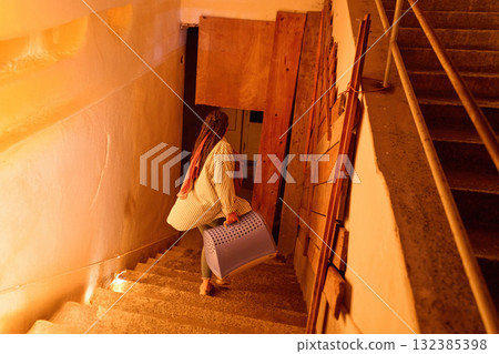 Woman carrying pet carrier up stairs in dimly lit building, interior shot, walking away, urban lifestyle, travel with pets, pet relocation, moving concept, apartment living. Woman carrying pet carrier up stairs in dimly lit building, interior shot, walking away, urban lifestyle, travel with pets, pet relocation, moving concept, apartment living. 132385398