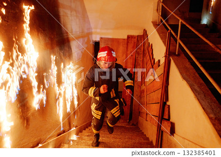 Firefighter running down burning stairs in emergency, wearing protective gear, helmet, and uniform, fire safety and rescue operation in dangerous situation, action scene 132385401