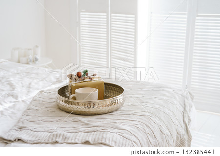 Christmas gift and cup on tray with lights on cozy white bed near decorated festive Christmas tree 132385441