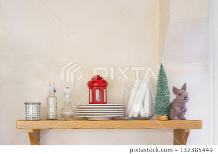 Wooden shelf with candles, red lantern, glass bottles, plates, and decorative Christmas bunny 132385449