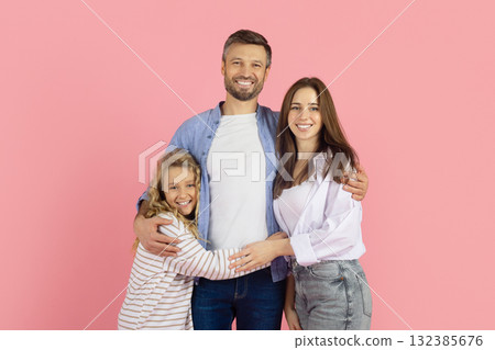 Smiling family standing close together in studio, pink background. Parents hugging daughter 132385676