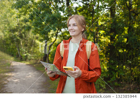 Smiling young woman with map and smartphone exploring nature trail Smiling young woman with map and smartphone exploring nature trail 132385944