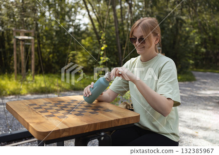 Young woman in sunglasses sitting outdoors opening reusable water bottle at picnic table 132385967