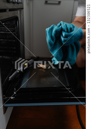 Vertical cropped shot of housekeeper male in gloves squeezing out wet rag after cleaning inside of gas oven with steam cleaner, close-up. Concept of hygiene and cleanliness in domestic environment. Vertical cropped shot of housekeeper male in gloves squeezing out wet rag after cleaning inside of gas oven with steam cleaner, close-up. Concept of hygiene and cleanliness in domestic environment. 132386521