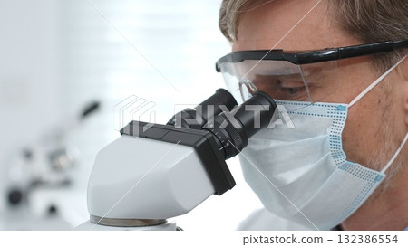 Close up of a scientist researching with a microscope, wearing protective glasses and a mask in a laboratory setting. Medicine, healthcare and science concept Close up of a scientist researching with a microscope, wearing protective glasses and a mask in a laboratory setting. Medicine, healthcare and science concept 132386554