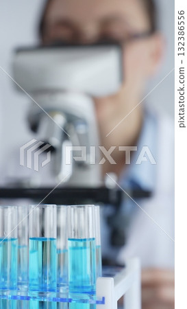 Close up of test tubes with blue liquid while scientist focusing on analyzing samples under a microscope in a laboratory setting. Medicine, healthcare and science concept 132386556