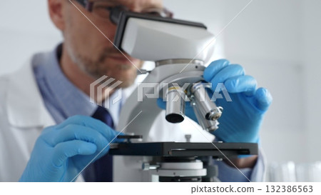Close up of male scientist in white coat and blue gloves adjusting microscope, conducting pharmaceutical research in modern laboratory. Medicine and science concept 132386563