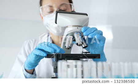 Woman science researcher wearing protective gear is checking microscope, analyzing blue test tubes in modern laboratory research setting. Medicine, and science concept Woman science researcher wearing protective gear is checking microscope, analyzing blue test tubes in modern laboratory research setting. Medicine, and science concept 132386580