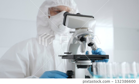Male scientific researcher or virologist wearing protective gear adjusting microscope, analyzing lab samples with precision in sterile research environment. Medicine concept 132386600