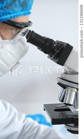 Medical scientist senior man wearing protective mask, glasses and bouffant cap using microscope doing scientific research in a laboratory. Medicine and science concept Medical scientist senior man wearing protective mask, glasses and bouffant cap using microscope doing scientific research in a laboratory. Medicine and science concept 132386606