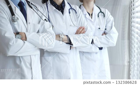Doctors in white uniforms with stethoscopes stand together with folded hands, symbolizing unity, competence, experience, and dedication to the healthcare profession 132386628