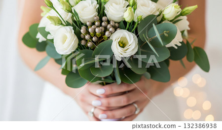 A hyper-realistic, high-detail close-up photograph of a bride's hands gently holding a gorgeous wedding bouquet filled with white Lisianthus, Silver Brunia berries, dominant Eucalyptus greenery. 132386938