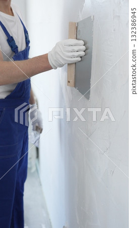 Man construction worker wearing protective gloves and blue construction coveralls, spreading plaster smoothly across wall using professional drywall taping knife Man construction worker wearing protective gloves and blue construction coveralls, spreading plaster smoothly across wall using professional drywall taping knife 132386945