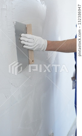 Close up of the construction worker hand wearing protective gloves, spreading plaster smoothly across wall using professional drywall taping knife realizing renovation project, vertical view 132386947