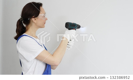 Professional female worker wearing blue overall, safety glasses and gloves, is using cordless drill to make hole in white wall during renovation work. Portrait view Professional female worker wearing blue overall, safety glasses and gloves, is using cordless drill to make hole in white wall during renovation work. Portrait view 132386952
