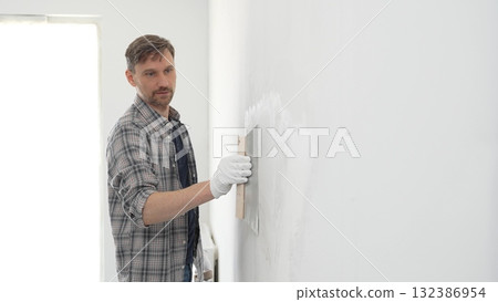 Male construction worker wearing protective gloves and plaid shirt, spreading plaster smoothly across wall surface using professional drywall taping knife during renovation project. Portrait view 132386954