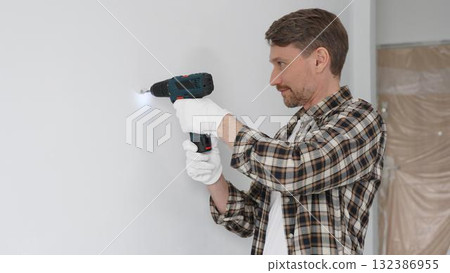 Male construction worker in checked shirt and safety gloves is screwing a screw into the wall using a cordless screwdriver, carrying out a careful home renovation. Portrait view Male construction worker in checked shirt and safety gloves is screwing a screw into the wall using a cordless screwdriver, carrying out a careful home renovation. Portrait view 132386955