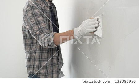 Unknown construction worker man wearing gloves and plaid shirt is using a putty knife to apply plaster on a white wall, during a home renovation or construction project Unknown construction worker man wearing gloves and plaid shirt is using a putty knife to apply plaster on a white wall, during a home renovation or construction project 132386956