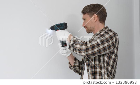 Male construction worker in checked shirt and safety gloves is screwing a screw into the wall using a cordless screwdriver, carrying out a careful home renovation. Portrait view 132386958