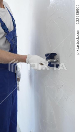 Professional construction worker wearing blue work overall is using a finishing trowel to carefully apply plaster on a wall, demonstrating expertise in home renovation Professional construction worker wearing blue work overall is using a finishing trowel to carefully apply plaster on a wall, demonstrating expertise in home renovation 132386963