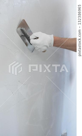 Professional construction worker is using a finishing trowel to carefully apply plaster on a wall, demonstrating expertise in home renovation and interior finishing, close-up of hand 132386965