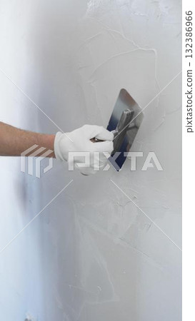 Professional construction worker is using a finishing trowel to carefully apply plaster on a wall, demonstrating expertise in home renovation and interior finishing, close-up of hand 132386966