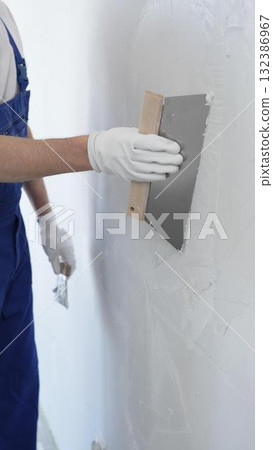 Man construction worker wearing protective gloves and blue construction coveralls, spreading plaster smoothly across wall using professional drywall taping knife Man construction worker wearing protective gloves and blue construction coveralls, spreading plaster smoothly across wall using professional drywall taping knife 132386967