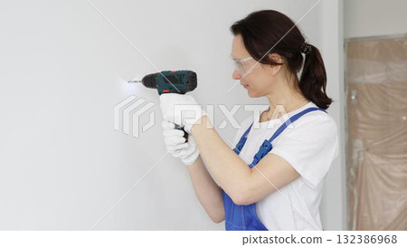 Professional female worker wearing blue overall, safety glasses and gloves, is using cordless drill to make hole in white wall during renovation work. Portrait view Professional female worker wearing blue overall, safety glasses and gloves, is using cordless drill to make hole in white wall during renovation work. Portrait view 132386968