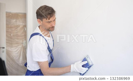 Man construction worker wearing protective white gloves and blue construction coveralls, is sanding white wall with blue hand block, preparing smooth surface during interior home renovation project Man construction worker wearing protective white gloves and blue construction coveralls, is sanding white wall with blue hand block, preparing smooth surface during interior home renovation project 132386969