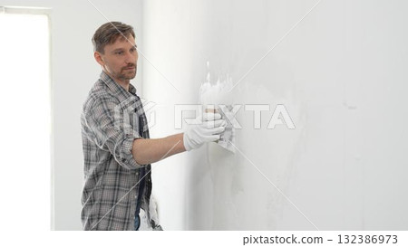 Construction worker spreading plaster on white wall, wearing protective gloves and t-shirt during home renovation project, working with professional skill and focus Construction worker spreading plaster on white wall, wearing protective gloves and t-shirt during home renovation project, working with professional skill and focus 132386973