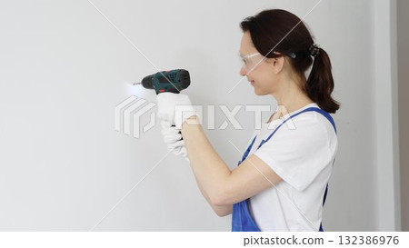 Professional female worker wearing blue overall, safety glasses and gloves, is using cordless drill to make hole in white wall during renovation work. Portrait view 132386976