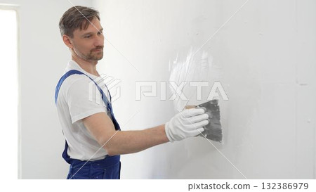 Middle aged male construction worker wearing gloves and blue overalls is using a putty knife to apply plaster on a white wall, performing home renovation 132386979