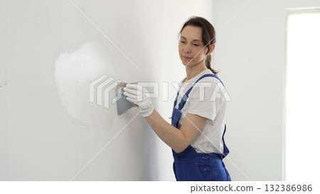 Construction worker woman spreading finishing putty on white wall, wearing protective gloves while smoothing surface with putty knife for precise home repair Construction worker woman spreading finishing putty on white wall, wearing protective gloves while smoothing surface with putty knife for precise home repair 132386986