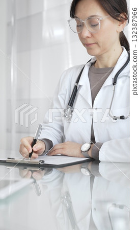 Female doctor in white coat and glasses writing medical notes on a clipboard at clinic desk, managing patient records. Medicine and health care 132387966