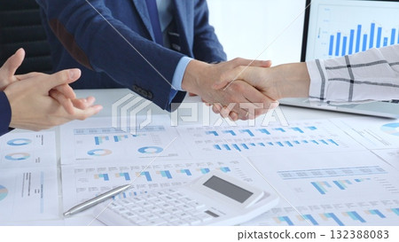 Business people shaking hands over financial documents and laptop, confirming successful partnership, agreement, or merger in a corporate office meeting 132388083
