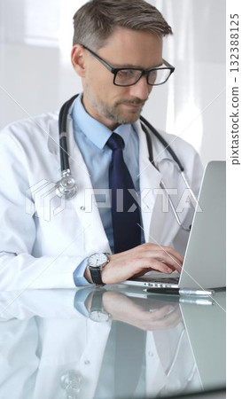 Male doctor in white coat with stethoscope, focused on laptop at reflective clinic desk, telemedicine consultation and research work. Medicine and health care 132388125