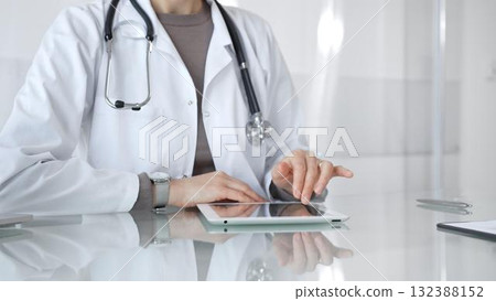 Physician in lab coat with stethoscope reviewing patient data on a tablet in a bright modern clinical office via touchscreen. Medicine and health care 132388152