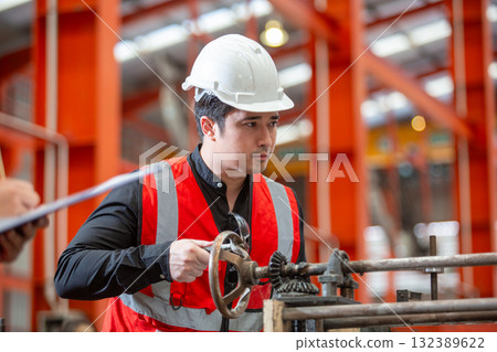 Industrial engineer wearing white safety helmet and red reflective vest operates heavy machinery in manufacturing plant, emphasizing focus, precision and hands on work in heavy factory. 132389622