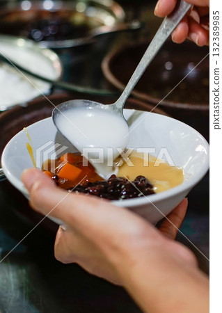 Close up of a hand pouring coconut milk over a bowl of traditional Indonesian sweet dessert made with black glutinous rice and sweet potato in palm sugar syrup	 132389985