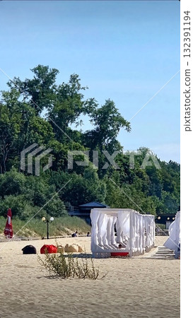 White wooden sun canopies with curtains dot the wide, sunny sand beach under a brilliant blue sky. White wooden sun canopies with curtains dot the wide, sunny sand beach under a brilliant blue sky. 132391194