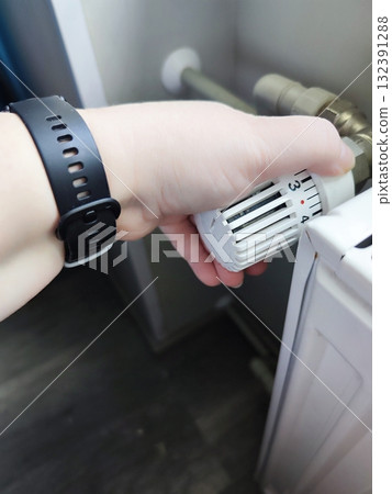 A person's hand, wearing a black wristwatch, is adjusting a white radiator valve. The valve displays numbers indicating temperature settings 132391288