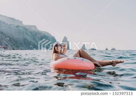 Woman, ocean, relaxation. Smiling woman floats on a pink donut ring enjoying summer vacation by rocky cliffs. 132397313